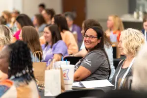 A smiling person wearing glasses and a "Book Nannies" shirt sits at a table with others during a gathering, surrounded by paper bags and notebooks.