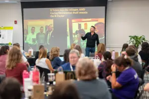 A speaker addresses an audience in a conference room, with a presentation screen displaying the message "It Takes ALL the villages."